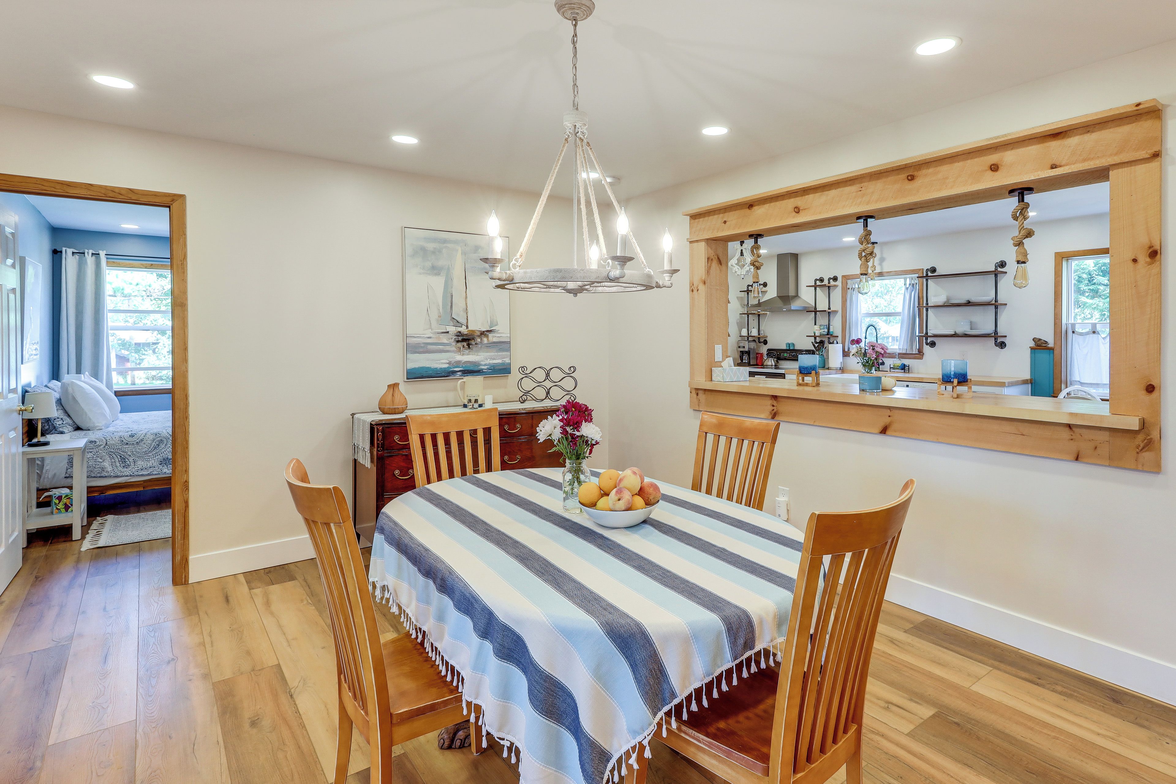 A dining area with a striped tablecloth, wooden chairs, a chandelier, and a kitchen visible through a large window.