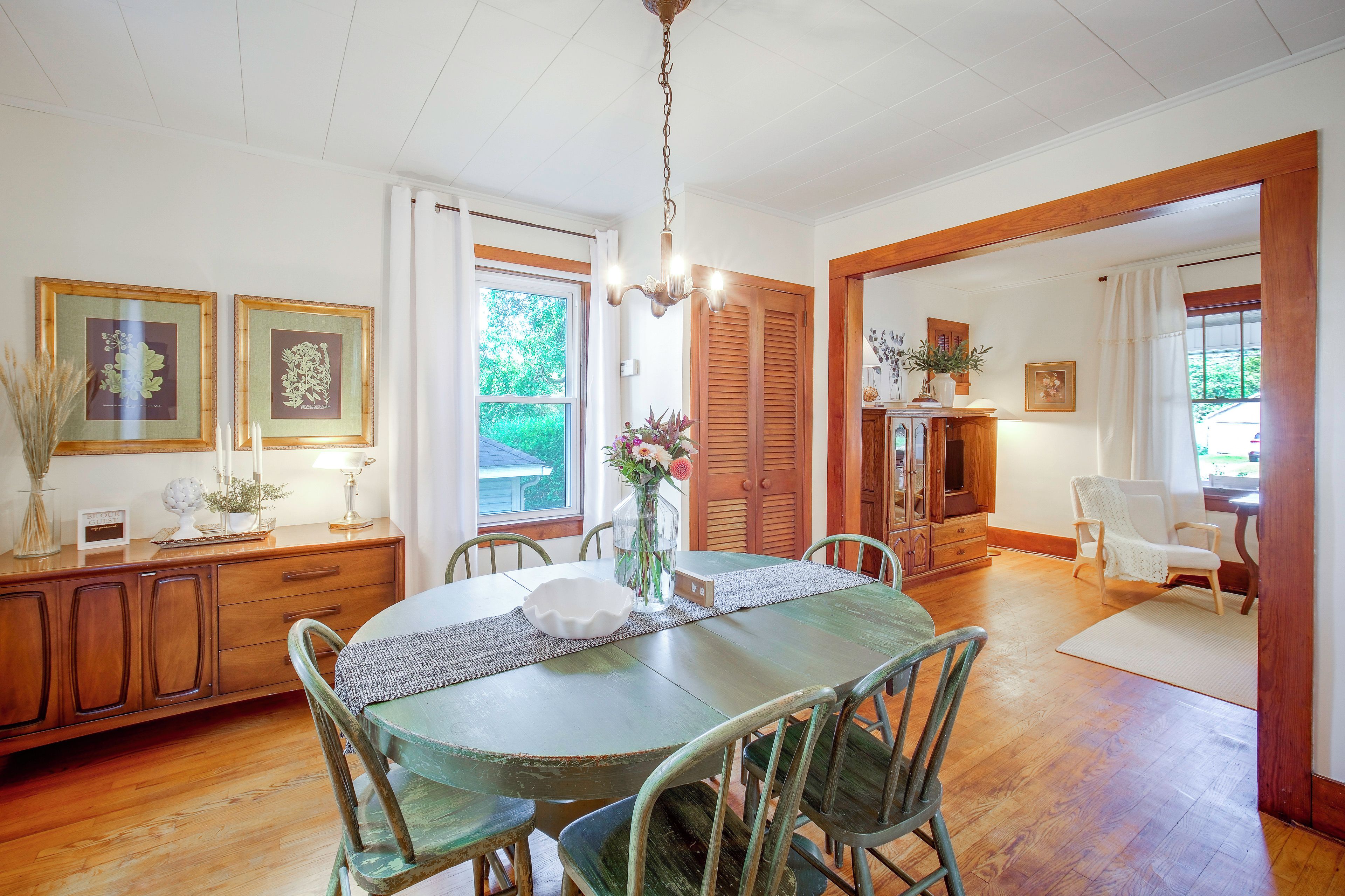 A dining area with a wooden table, chairs, and a vase with flowers. There are framed botanical prints on the wall, a wooden cabinet, and a door leading to another room.