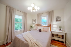 A bedroom with a bed, bedside tables, a nightstand, a lamp, a framed picture, and a window with curtains.
