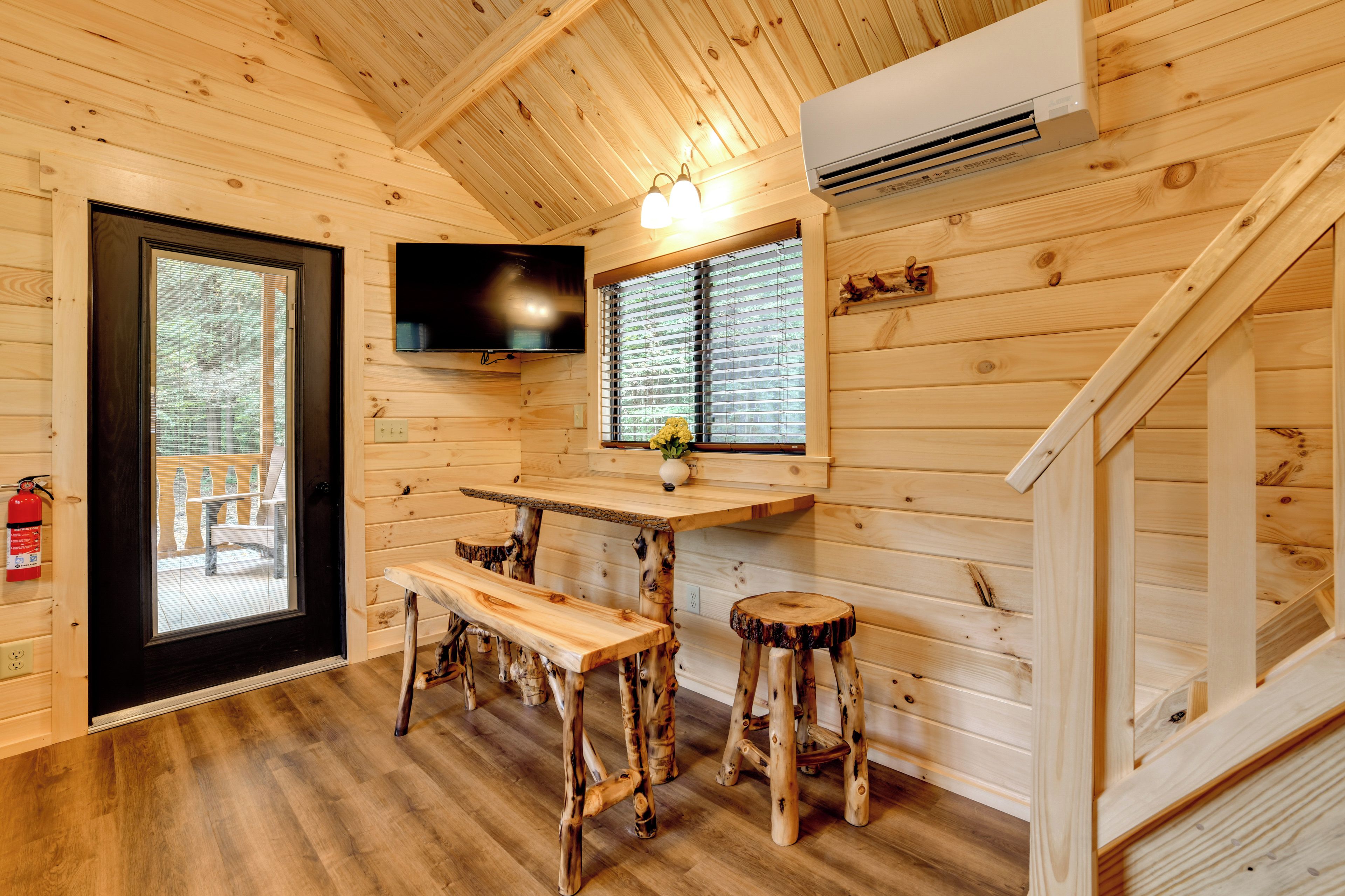 A wooden cabin interior with a television mounted on the wall, a dining table with benches, a window with blinds, and a staircase.