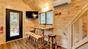 A wooden cabin interior with a television mounted on the wall, a dining table with benches, a window with blinds, and a staircase.