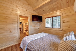 A wooden cabin bedroom with a bed, a TV mounted on the wall, and a window with a view of trees. - Hike, Bike & Stargaze! Oxford Cabin w/ Fire Pit (Oxford)