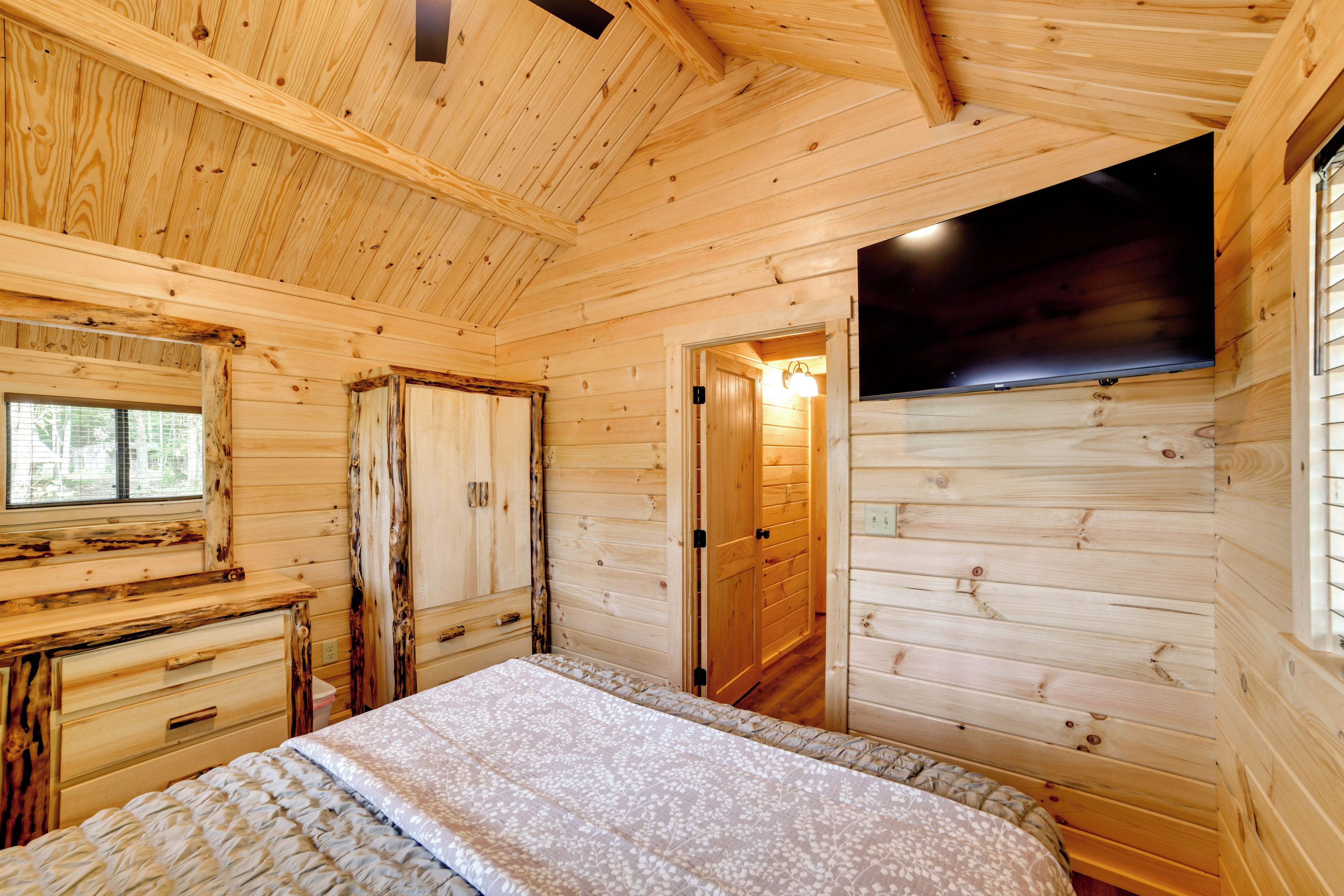 A wooden cabin room with a bed, a dresser, a television, and a door leading to another room.