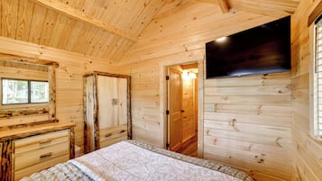 A wooden cabin room with a bed, a dresser, a television, and a door leading to another room.