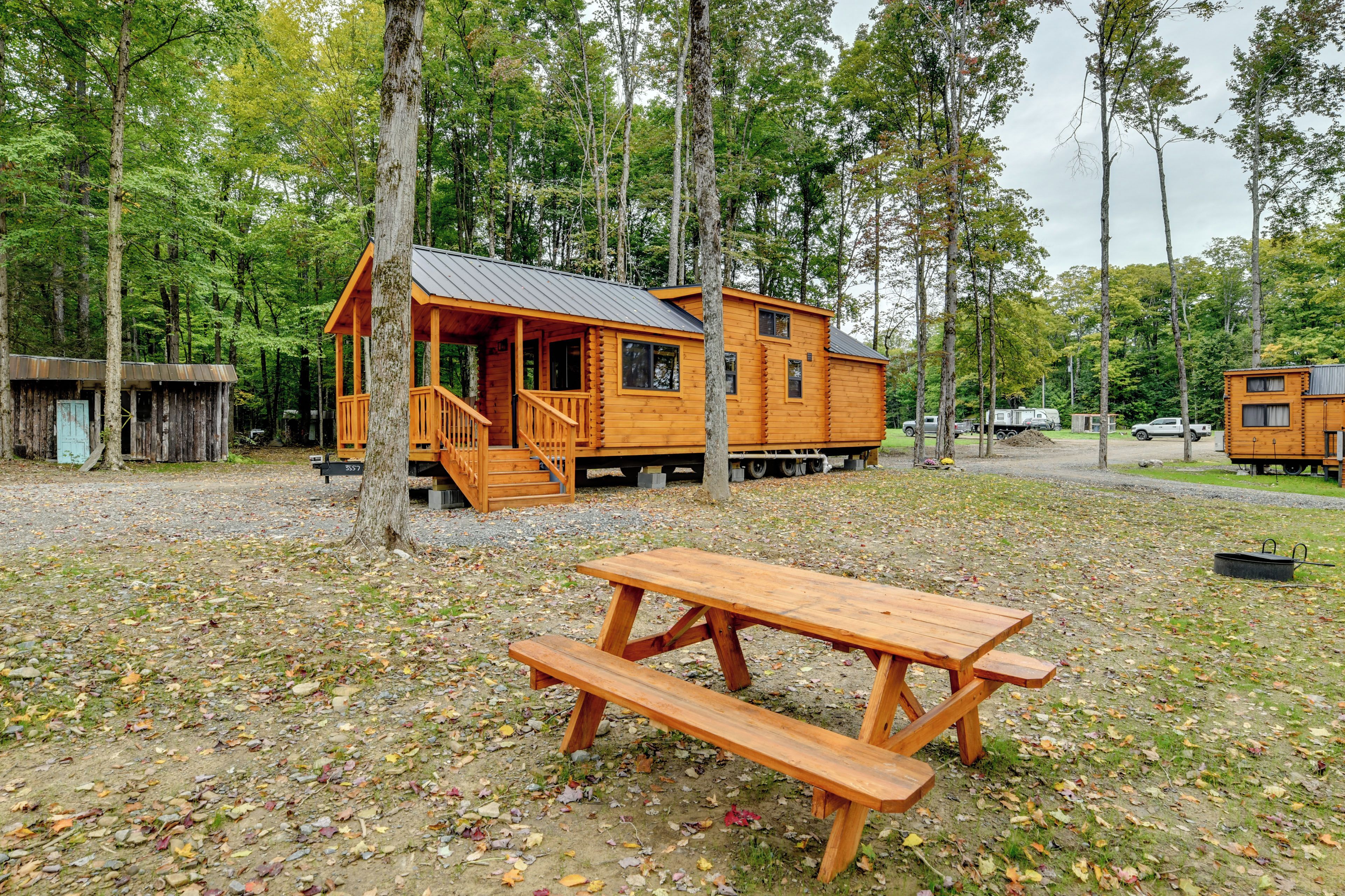 A rustic cabin with a porch, a picnic table, and a small shed in a wooded area.