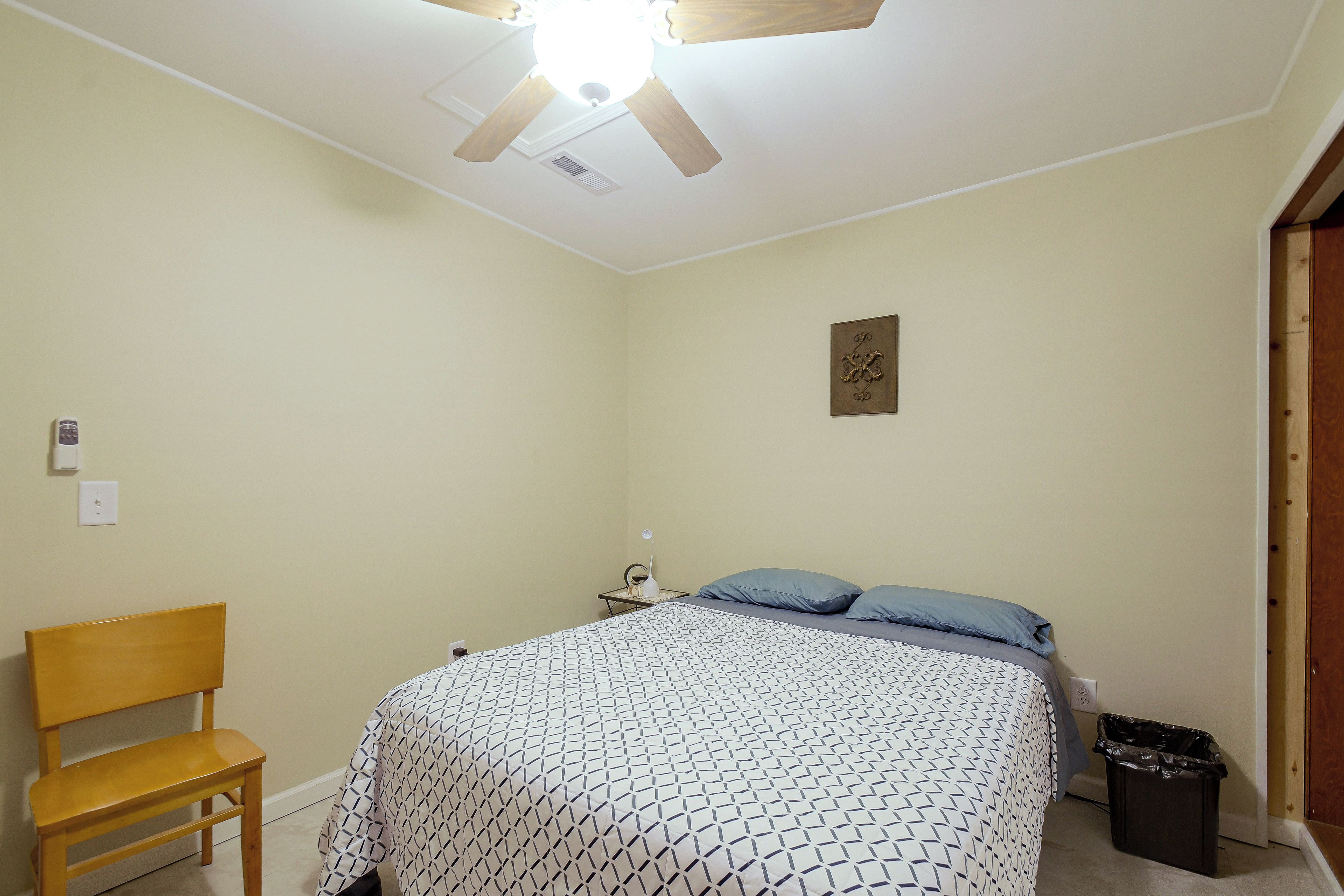 A bedroom with a bed, a chair, a bedside table, a ceiling fan, and a wall-mounted light fixture.