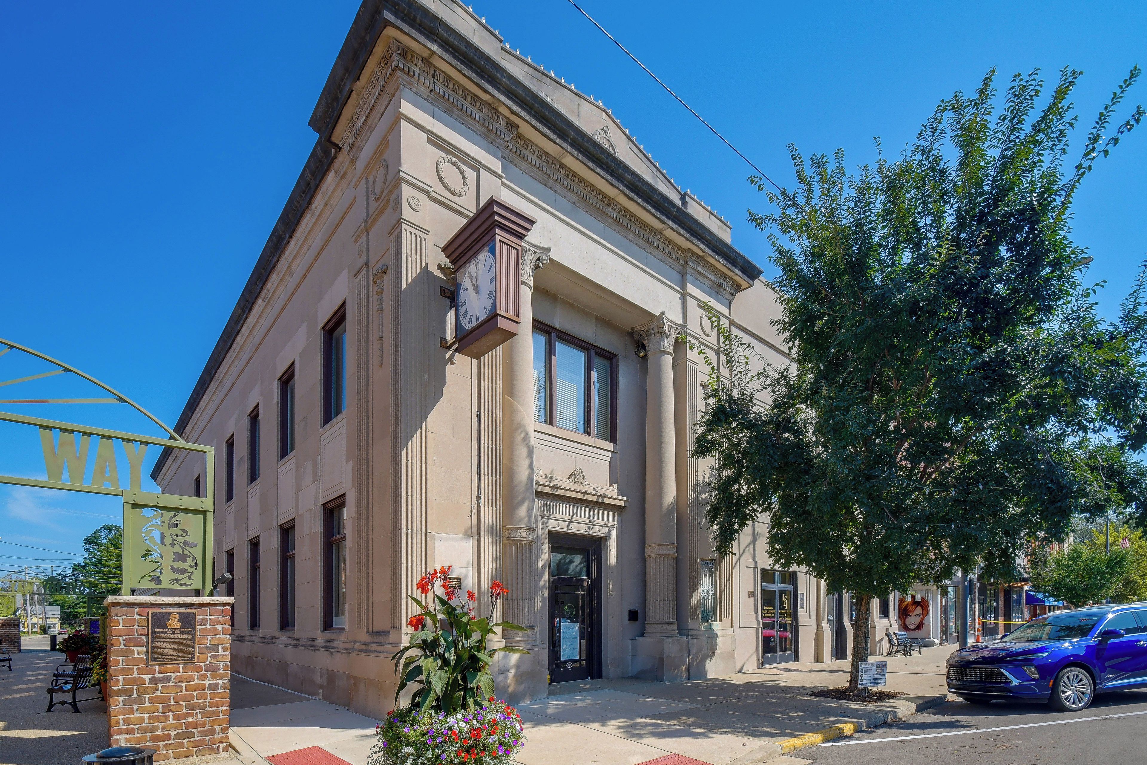 A classical building with a clock, a bench, and a tree in front.