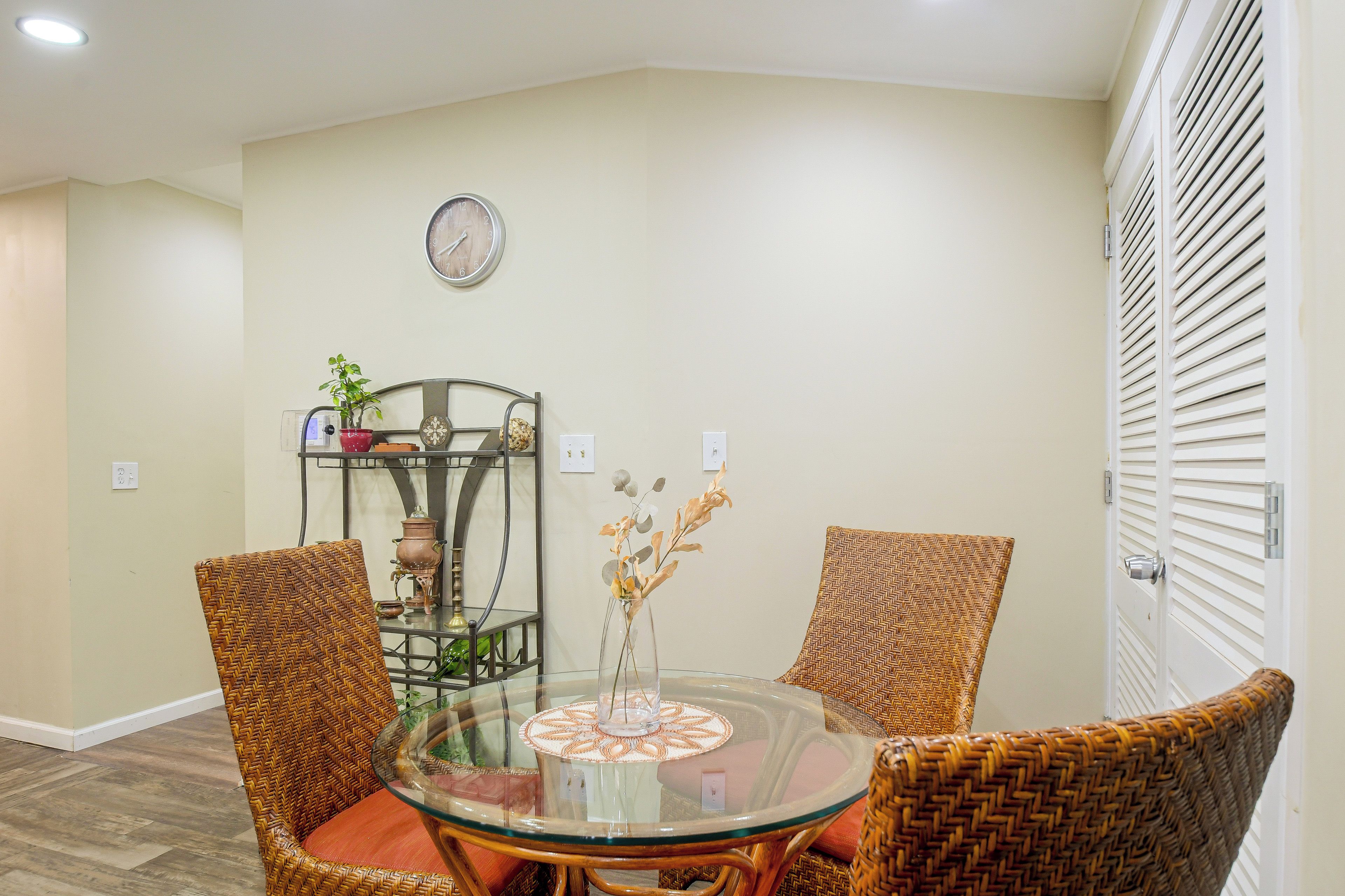 A dining area with a round glass table, wicker chairs, a wall clock, and a shelf with decorative items.