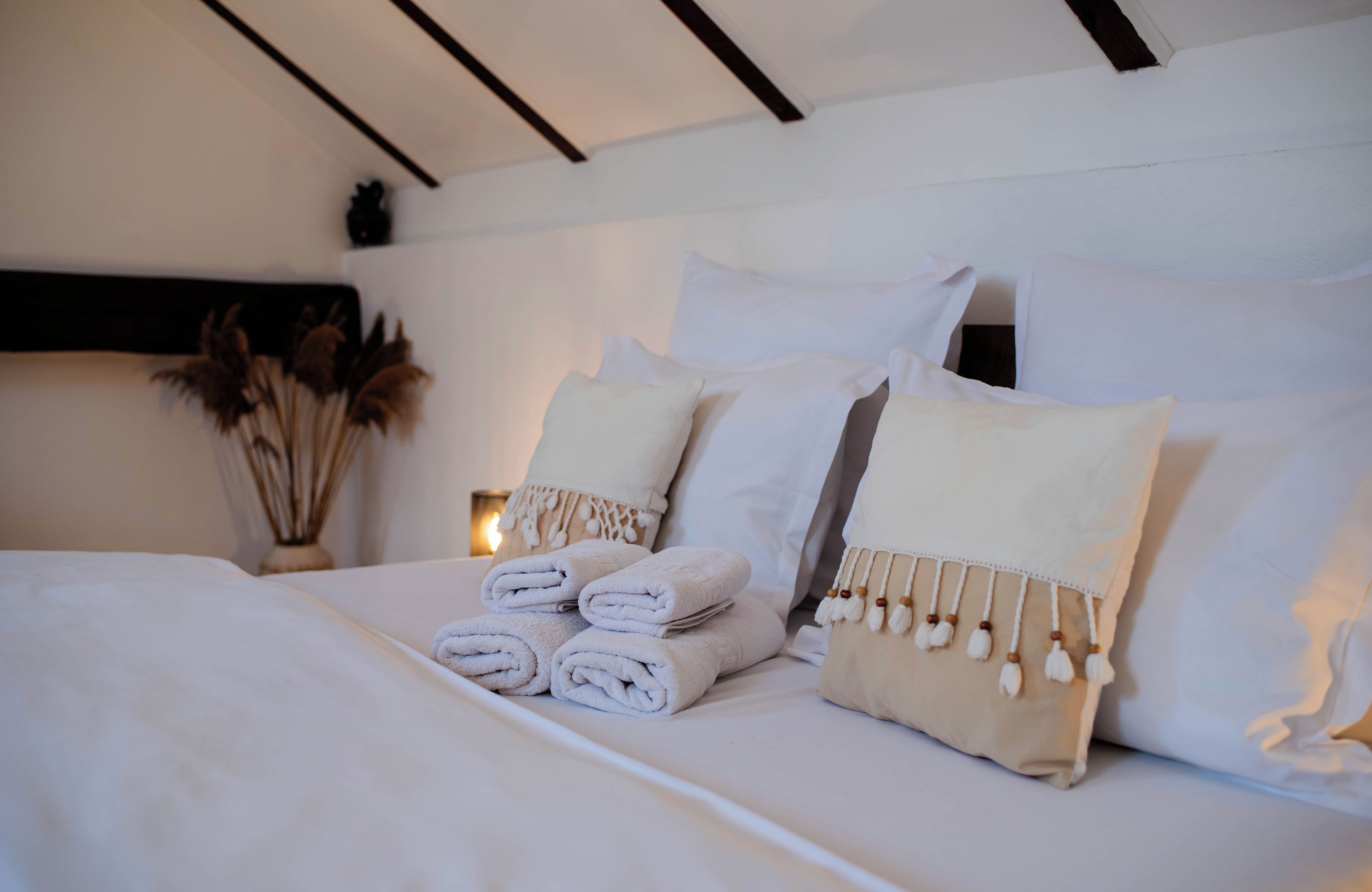 A neatly made bed with rolled towels, a decorative pillow with tassels, and a vase with dried plants.
