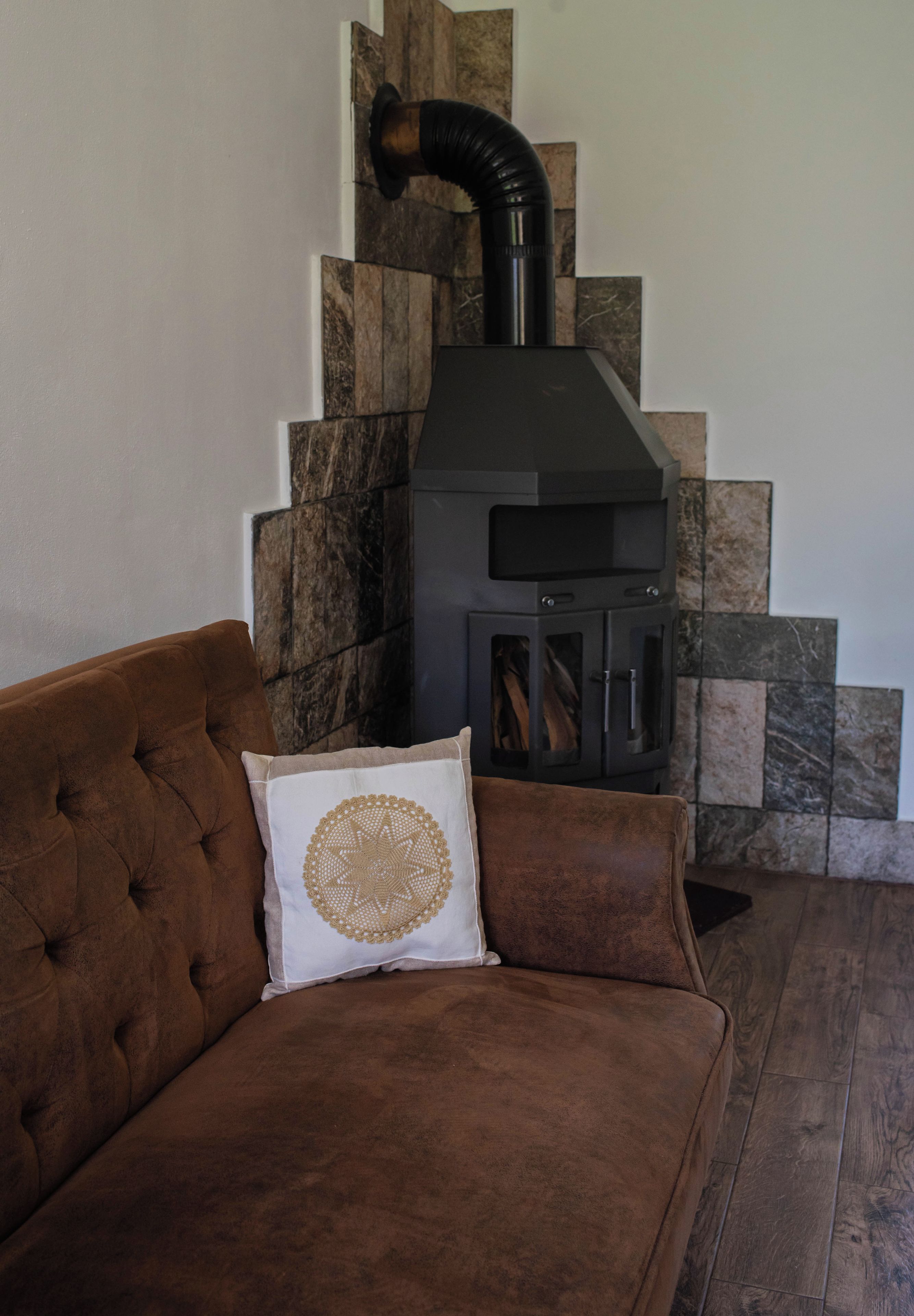 A living room with a brown sofa, a white pillow with a gold pattern, and a black wood-burning stove against a stone wall.