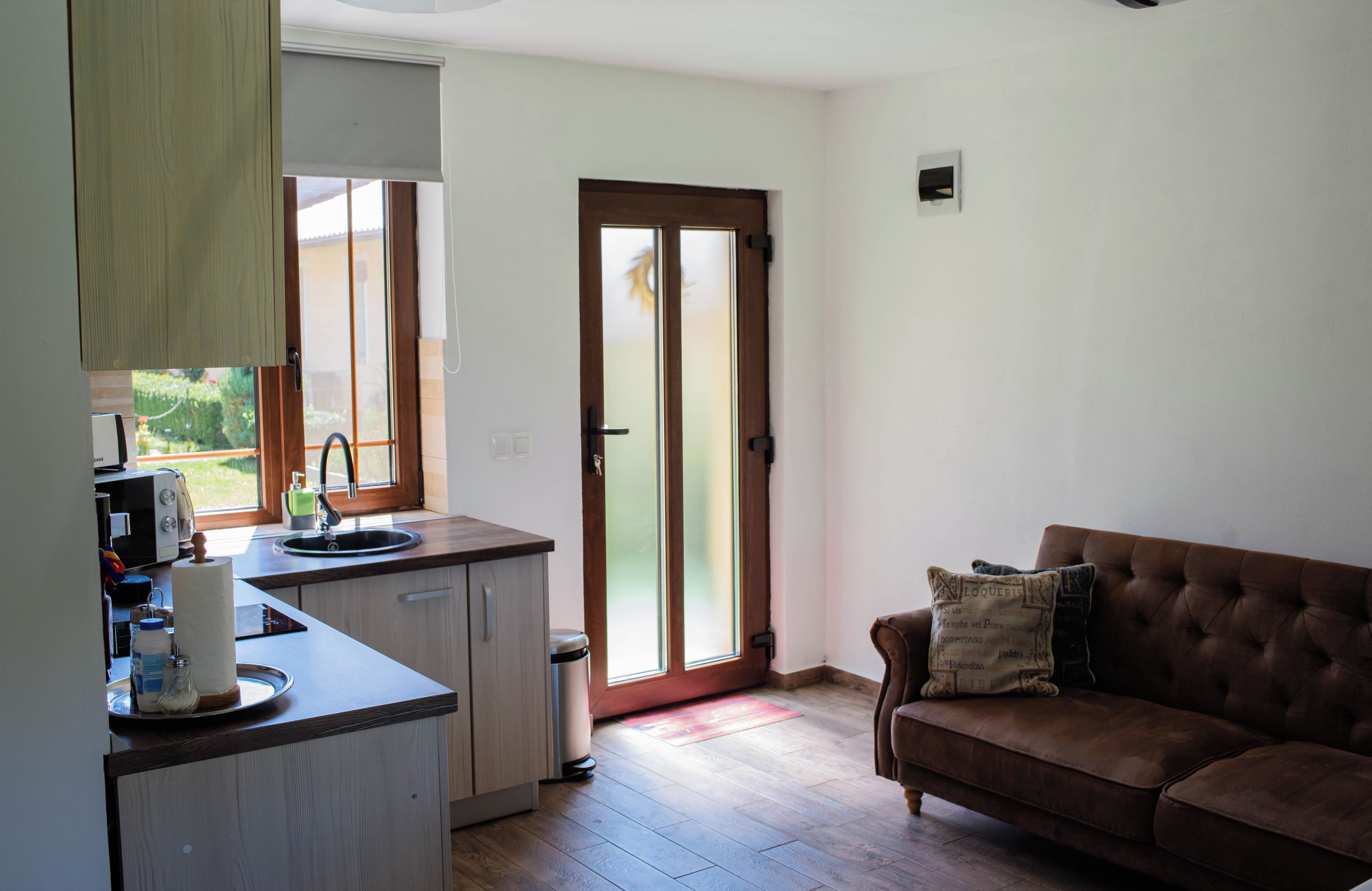 A compact living space with a kitchenette, a brown leather sofa, and a sliding glass door leading to a garden.