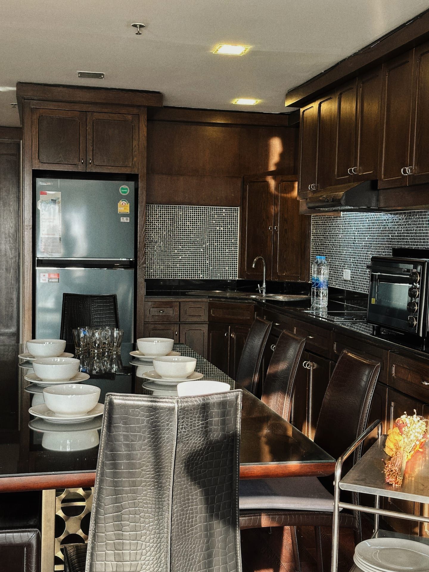 A modern kitchen with dark wood cabinets, a stainless steel refrigerator, a microwave, and a dining area with a long table and chairs.