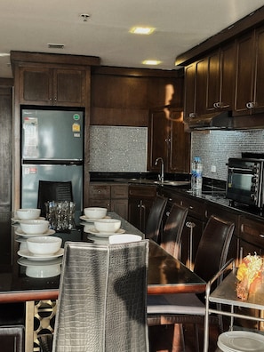 A modern kitchen with dark wood cabinets, a stainless steel refrigerator, a microwave, and a dining area with a long table and chairs. - Spacious 125 sqm Executive 3-Bedroom Residence in Silom-Sathon (Krung Thep Maha Nakhon)