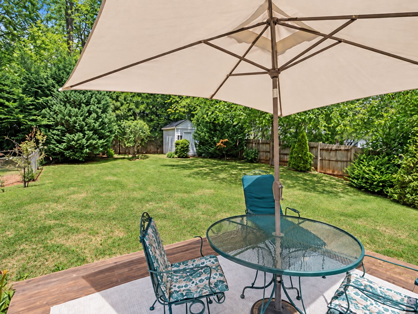 A backyard patio with a glass table and chairs, an umbrella, and a wooden deck.