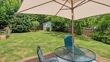A backyard patio with a glass table and chairs, an umbrella, and a wooden deck.
