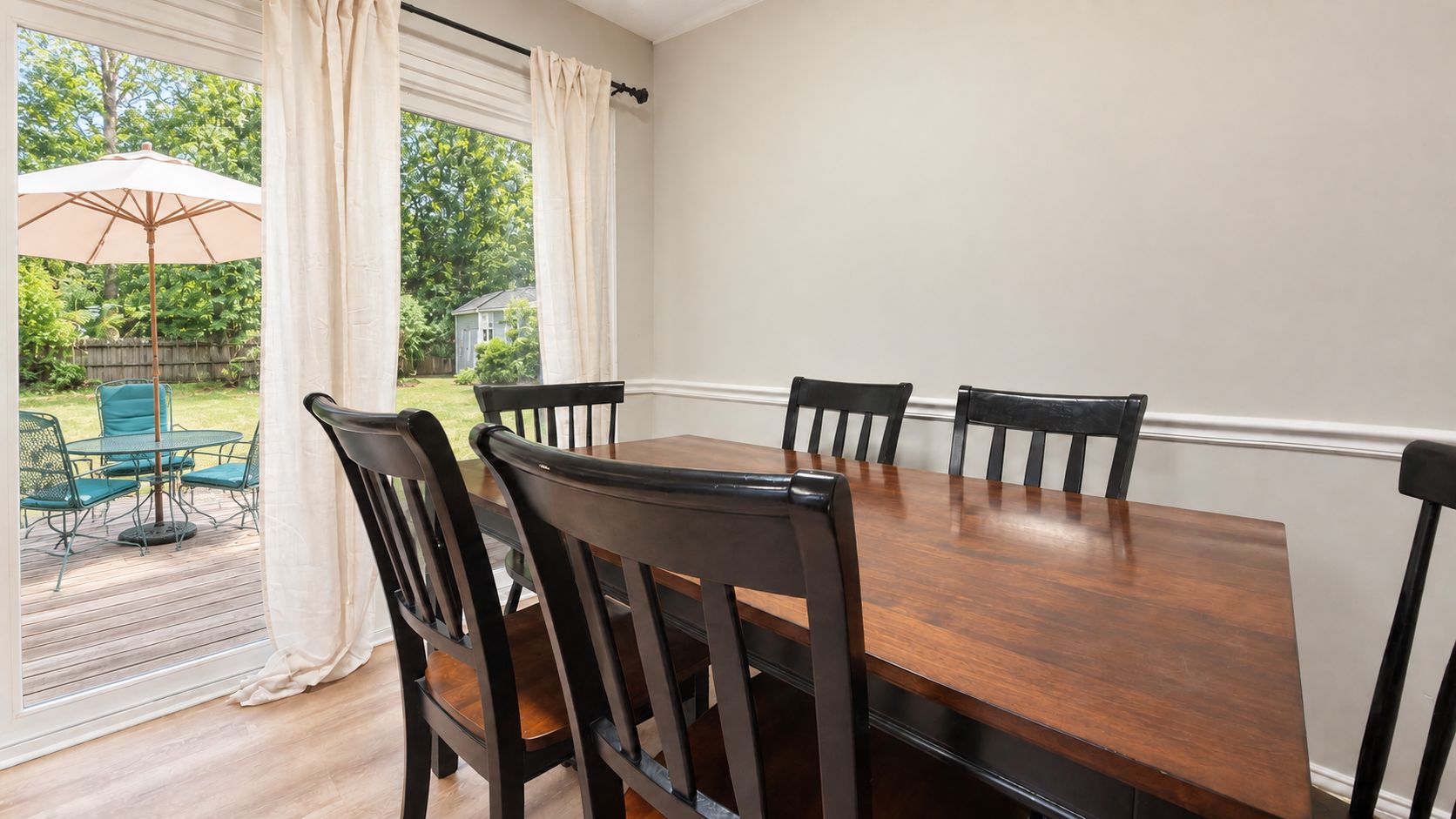 Uma área de refeições com mesa e cadeiras de madeira, janela com cortinas e vista para um jardim.