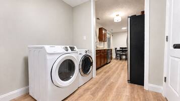 A laundry area with two front-loading washers and dryers, a wooden floor, and a kitchen in the background.