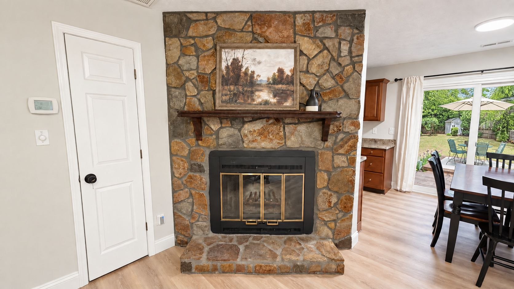 A stone fireplace with a mantel, a dining area with a table and chairs, and a view of the outdoors through a glass door.