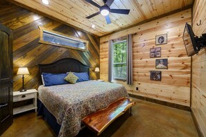 Ein Schlafzimmer mit Holzboden, einem Bett mit gemusterten Betttextilien, einer Bank, einem Nachttisch mit Lampe und einem Fenster mit Vorhängen.