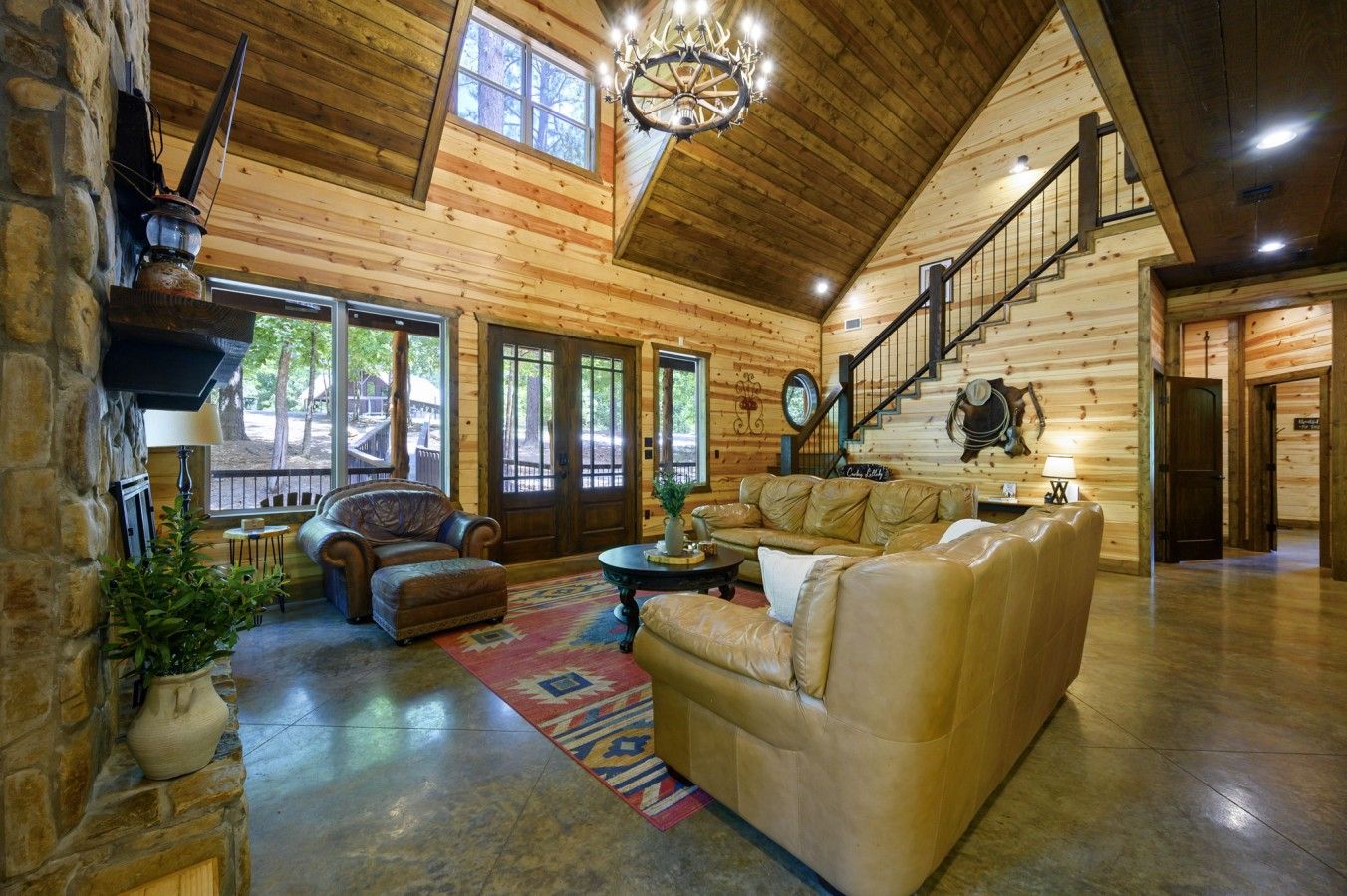 A spacious log cabin interior with a leather sofa, a fireplace, a staircase, and a large window overlooking a deck.