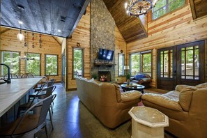 A log cabin interior with a stone fireplace, leather sofas, a dining area, and large windows. - Whispering Meadows|Fooseball|Firepit (Broken Bow)