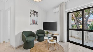 A modern living room with a large TV, two green armchairs, a small round table, and a view of the outdoors through a sliding glass door. - Falésia Stylish 2BR W/Balcony & Pool by LovelyStay (Olhos de Água)
