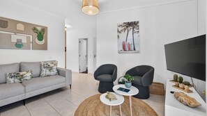 Un salón moderno con un sofá gris, dos sillones, una mesa de centro redonda, un televisor de pantalla plana, y una pared con arte abstracto y una escena de playa.