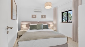 A bedroom with a bed, bedside lamps, an air conditioning unit, and a window with curtains. - Falésia Stylish 2BR W/Balcony & Pool by LovelyStay (Olhos de Água)