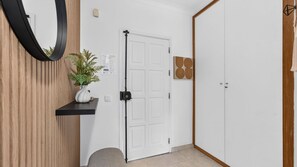 A hallway with a white door, a potted plant on a shelf, and a wooden wall panel. - Falésia Stylish 2BR W/Balcony & Pool by LovelyStay (Olhos de Água)