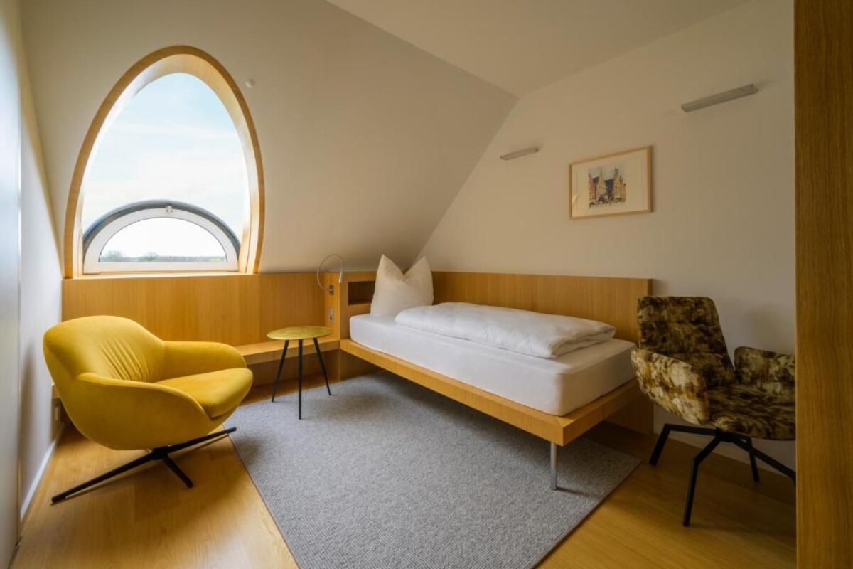 A room with a bed, a yellow chair, a patterned chair, a small table, and a large arched window.