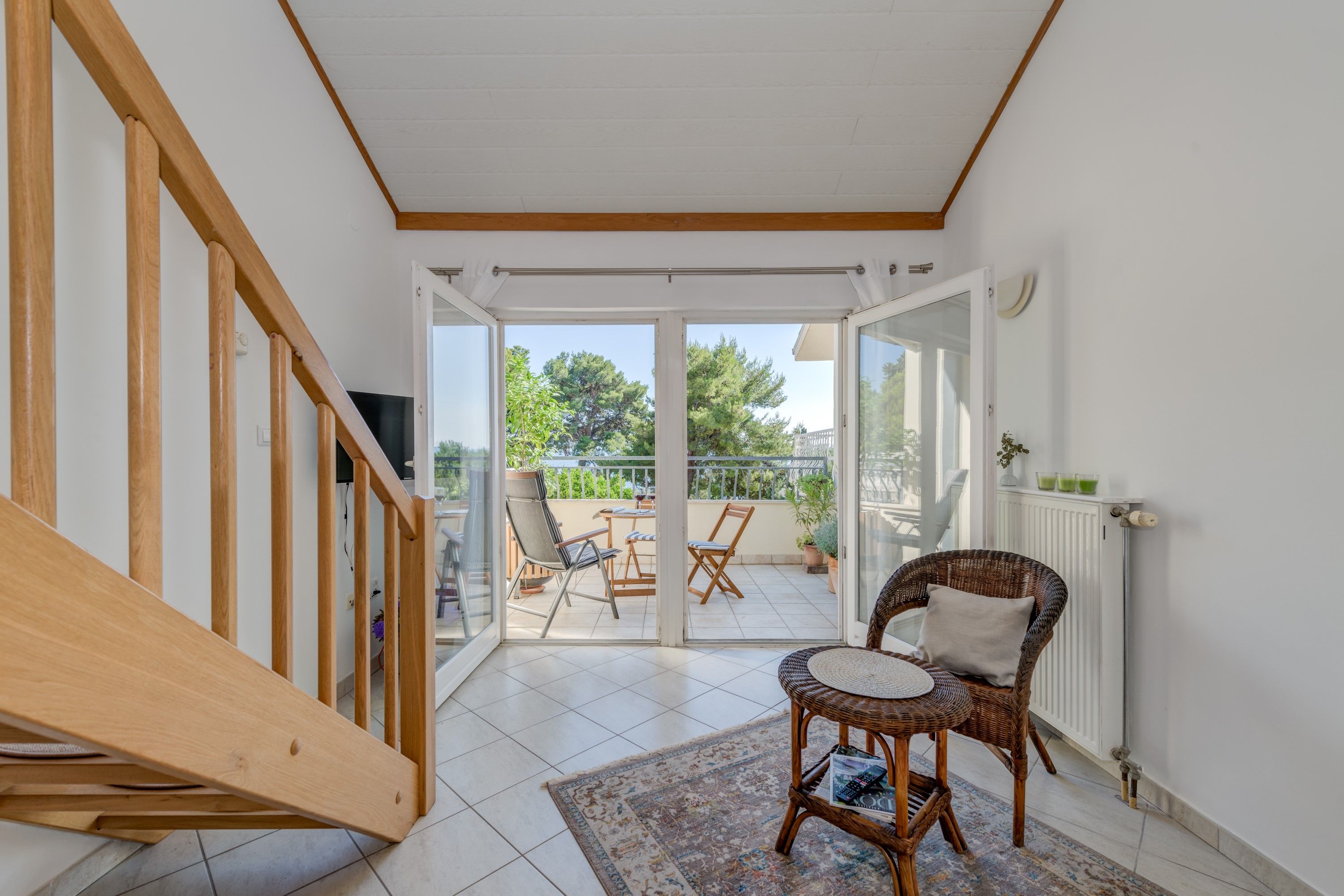 A room with a wooden staircase, a wicker chair, a small table, and a view of an outdoor balcony with chairs and plants.