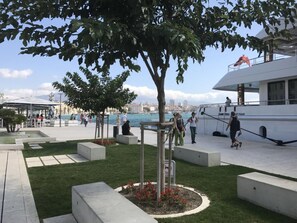 Un paseo marítimo con un gran árbol, zonas de estar y un barco blanco amarrado cerca.