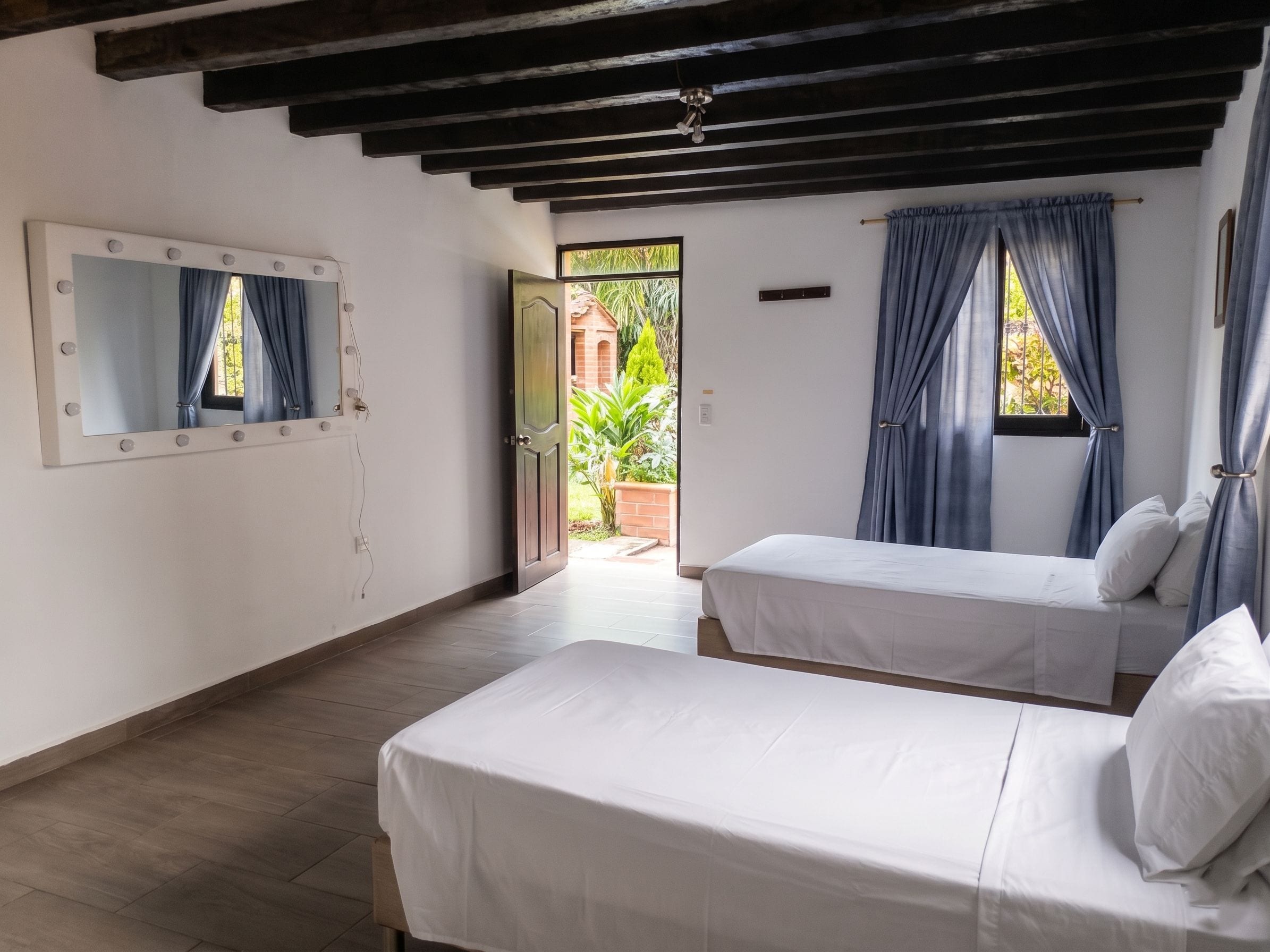 Una habitación con dos camas, un tocador con espejo e iluminación, y vistas al exterior con vegetación.