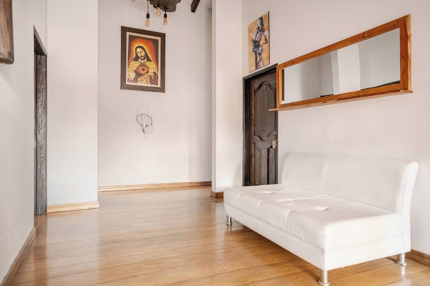 Una habitación con un sofá blanco, suelo de madera, una puerta de madera oscura y un espejo montado en la pared.