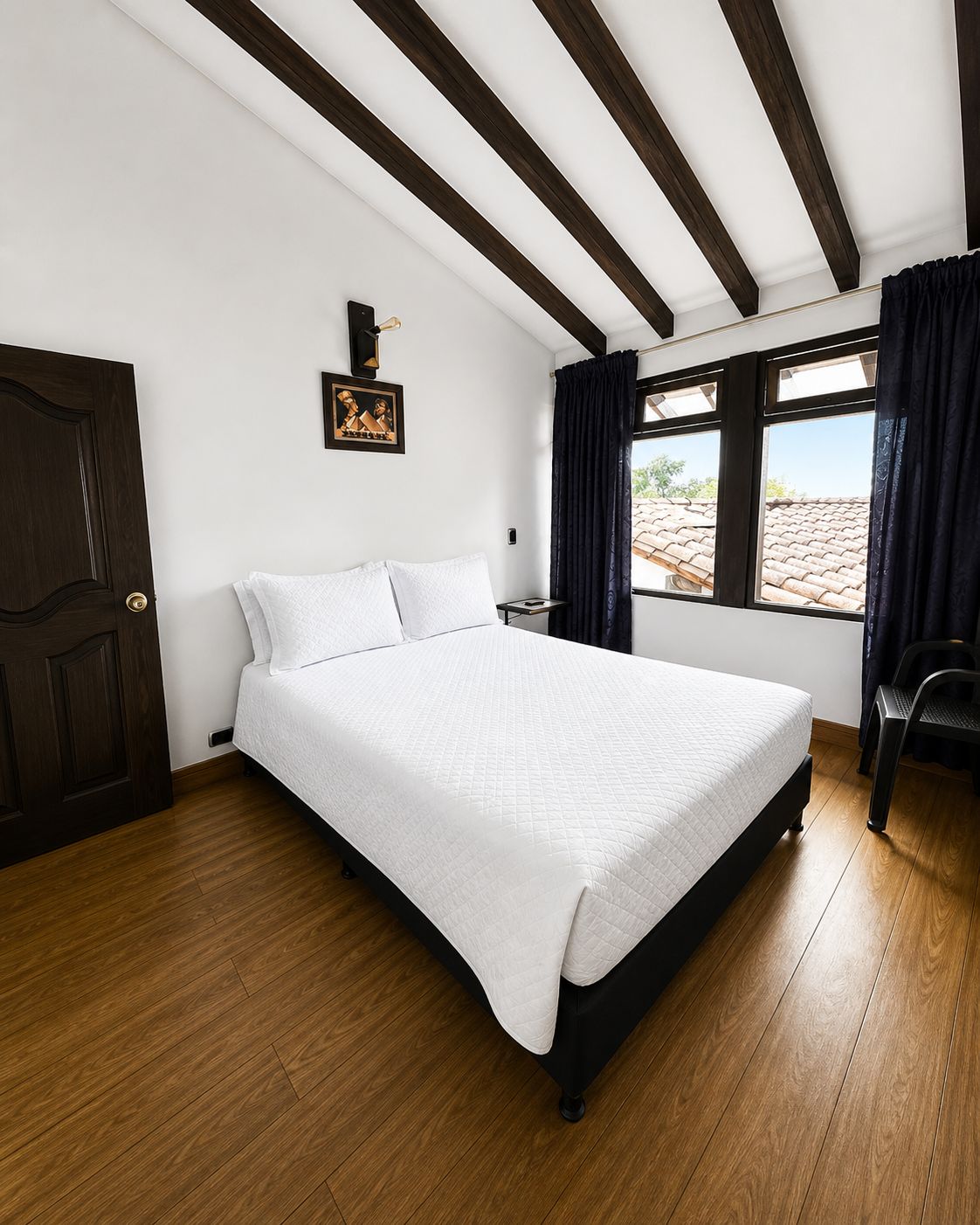 A hotel room with a large bed, wooden flooring, a dark door, a window with curtains, and a small bedside table.