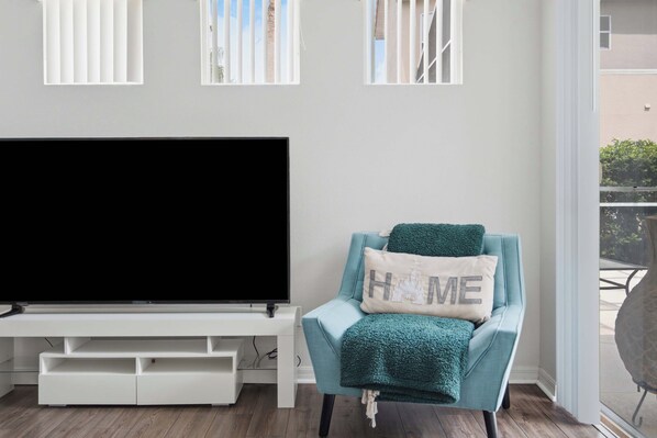 A living room with a flat-screen TV on a white stand, a teal armchair with a 'HOME' pillow, and a view of a balcony with a railing.