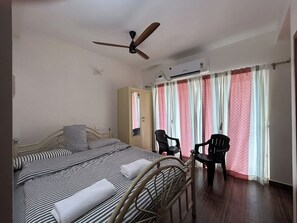 A hotel room with a bed, two chairs, a wardrobe, an air conditioning unit, and a ceiling fan.