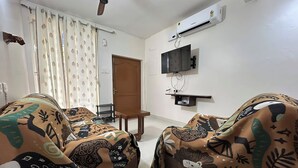 Ein Wohnzimmer mit zwei sofas, bezogen mit gemustertem Stoff, einem Deckenventilator, einer Klimaanlage, einem an der Wand befestigten Fernseher und einer Tür zu einem anderen Raum.