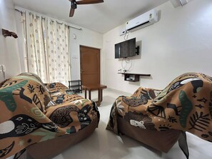 A living room with two sofas covered in patterned fabric, a ceiling fan, an air conditioning unit, a television on the wall, and a door leading to another room. - chennai beach villa (Kovalam)