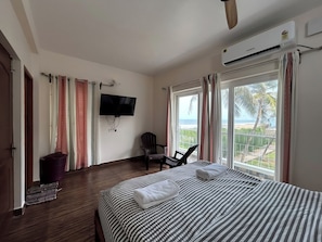 Una habitació d'hotel amb un llit, una cadira, una televisió i un balcó amb vista a la platja.