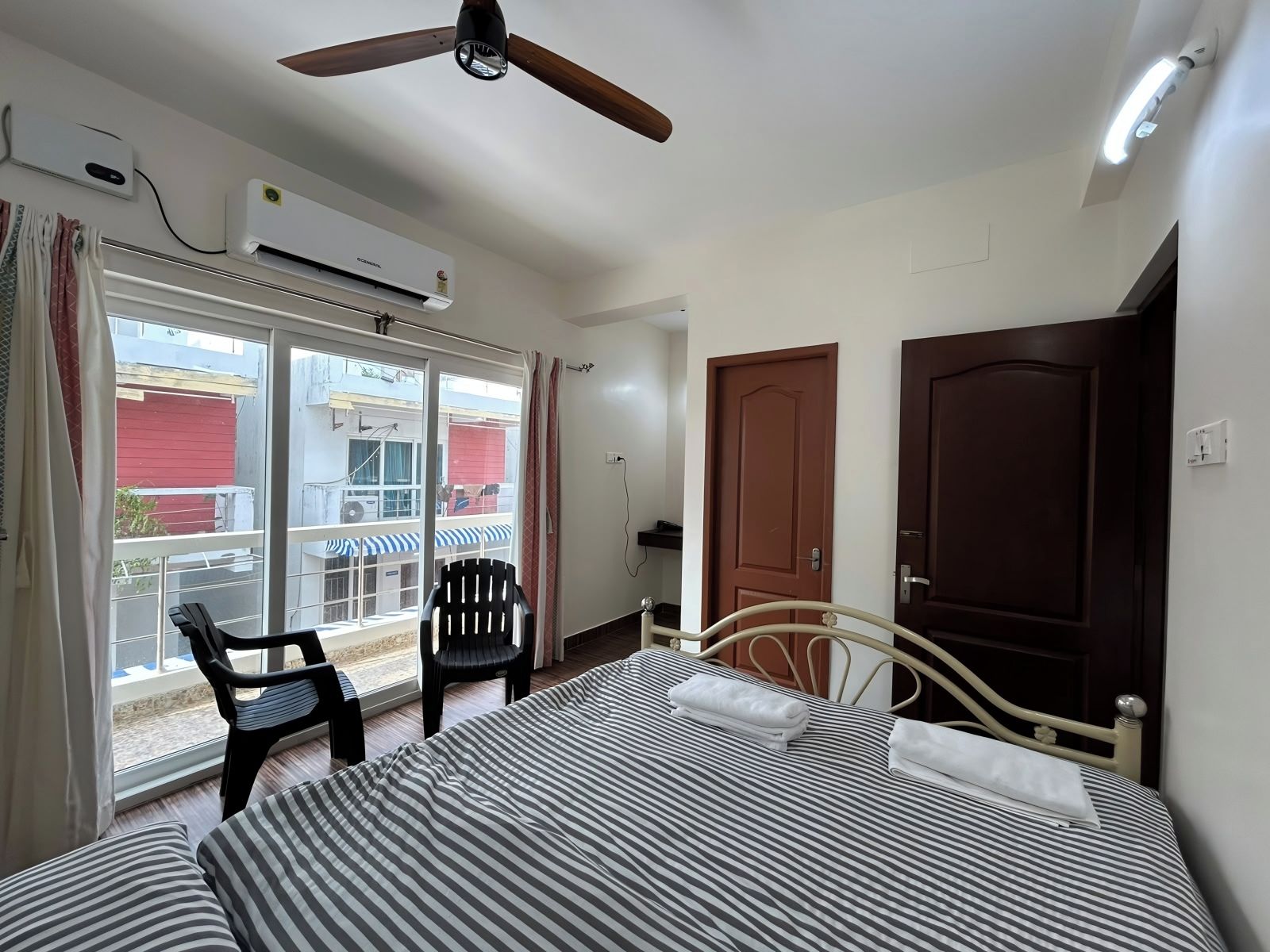 A hotel room with a bed, a chair, a ceiling fan, an air conditioning unit, and a view of the outside.