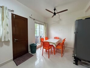 A room with a dining table, chairs, a ceiling fan, a window with bars, and a refrigerator. - chennai beach villa (Kovalam)