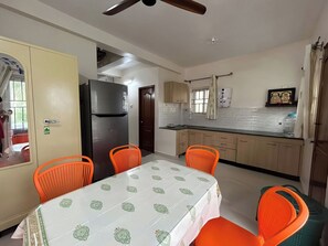 A compact kitchen with a refrigerator, a dining table with orange chairs, and a ceiling fan.