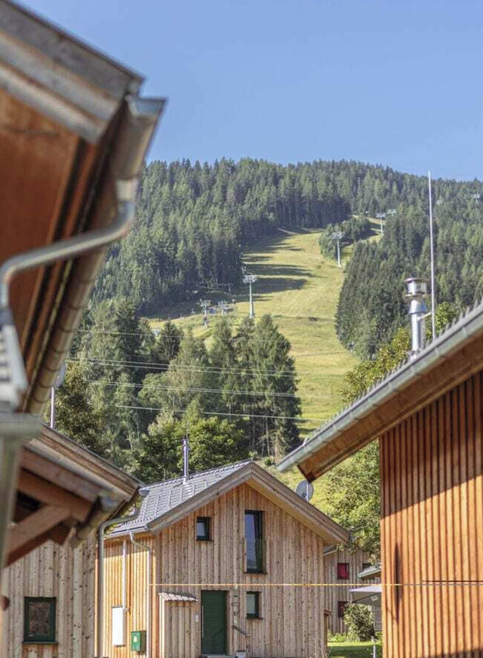 A wooden cabin with a view of a forested hill and a ski slope in the background.