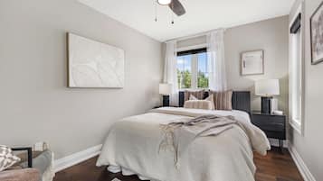 A bedroom with a bed, a ceiling fan, a window with curtains, a bedside table with a lamp, and a framed picture on the wall.