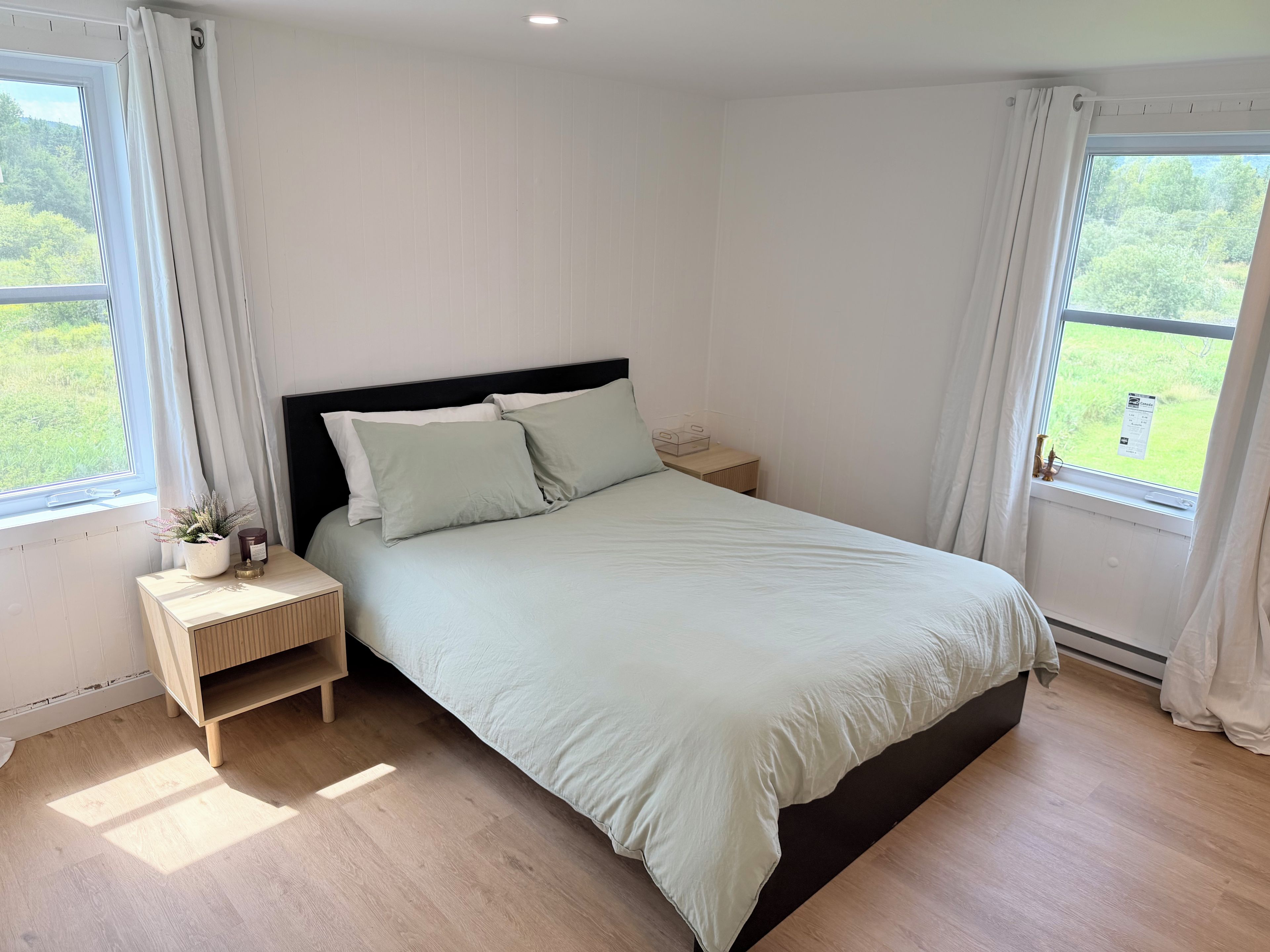 A bedroom with a bed, two bedside tables, a nightstand, a window with curtains, and a view of greenery outside.