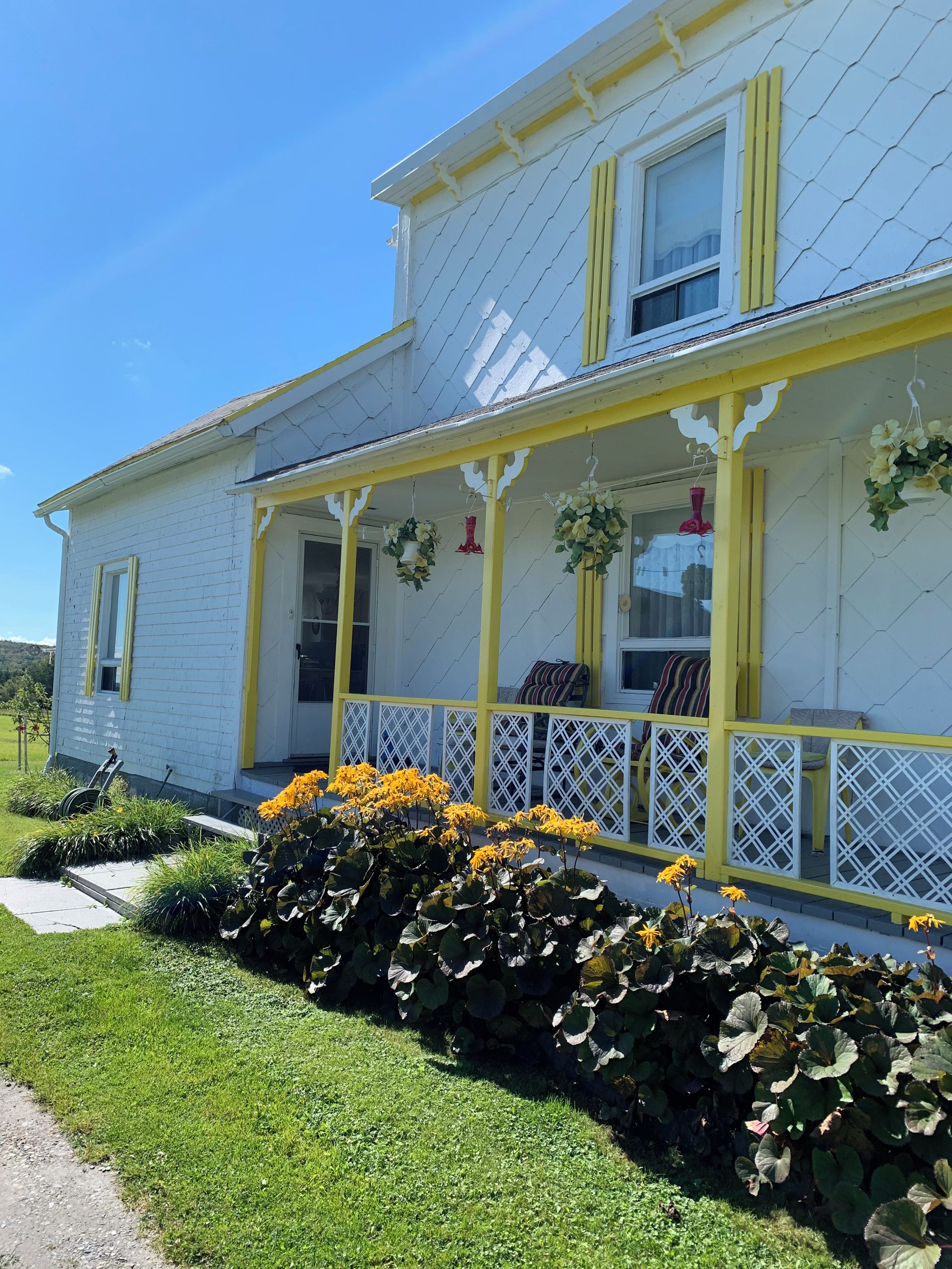 Una casa de dos pisos con un porche, detalles en amarillo y un pequeño jardín con flores.