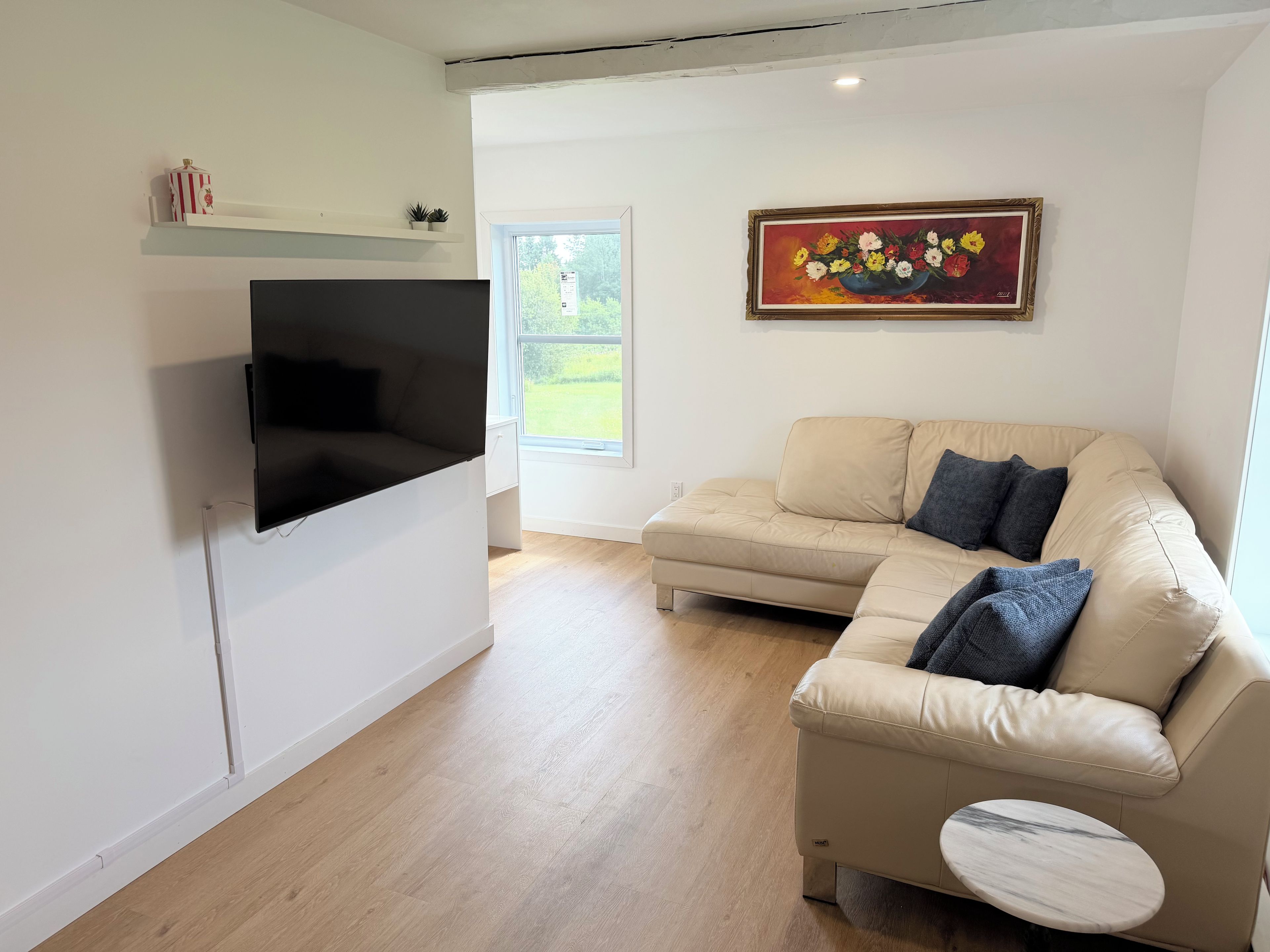 A modern living room with a flat-screen TV mounted on the wall, a beige sectional sofa with cushions, a small side table, and a painting of flowers on the wall.