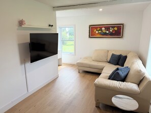 A modern living room with a flat-screen TV mounted on the wall, a beige sectional sofa with cushions, a small side table, and a painting of flowers on the wall. - Charming country cottage (Sainte-Sabine)