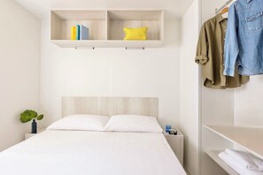 Ein Schlafzimmer mit einem Bett, einem Nachttisch, einem Regal mit Büchern, einem Kissen, einer aufgehängten Jacke und einer kleinen Pflanze.